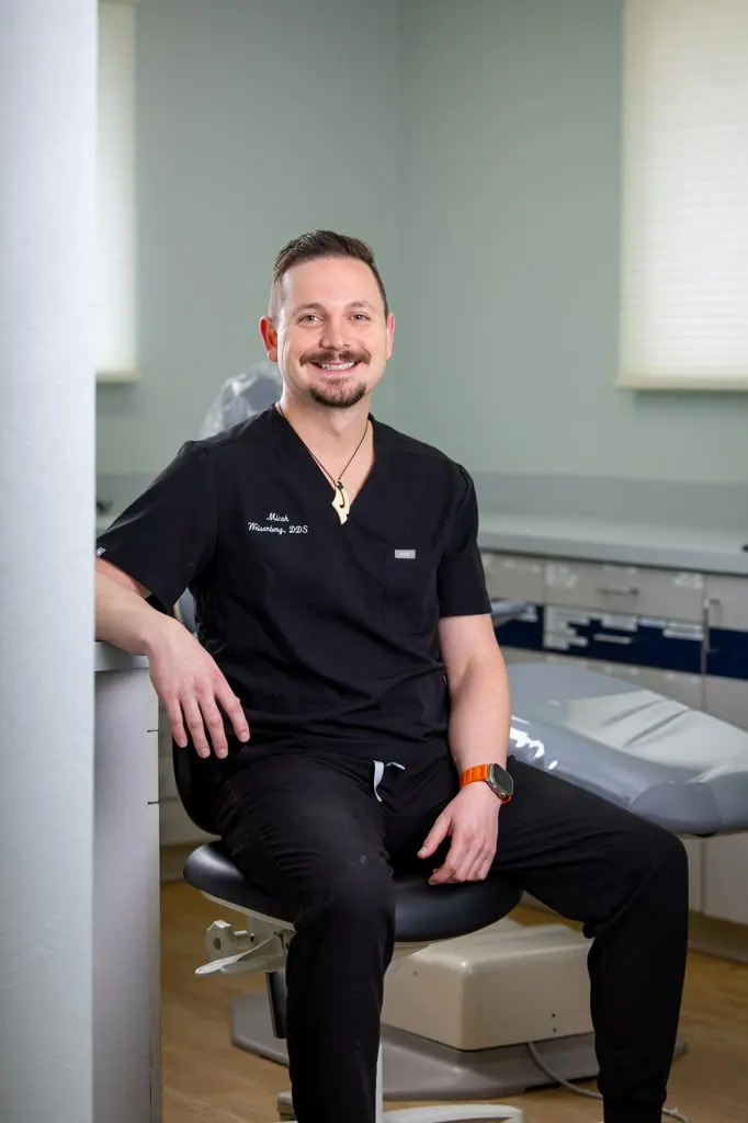 Dr. Micah Weisenberg in black scrubs, seated beside a dental chair in his operatory